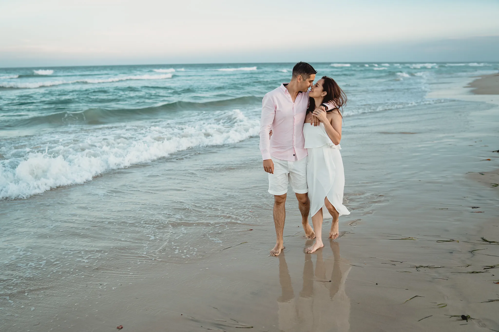 Rafaela and Mark walking arm in arm along the shoreline at blue hour, their reflections in the wet sand, photographed by Sean